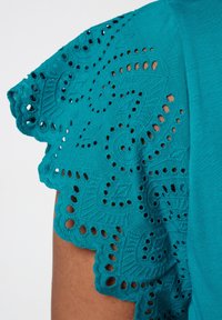 Top teal avec des manches en dentelle brodée avec des motifs découpés et des bords festonnés, contrastant avec un corps en tissu lisse.