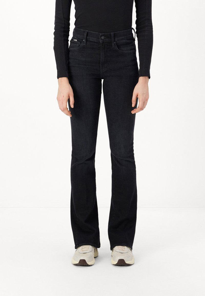 Black flared jeans with a high waist, made of denim. Features two front pockets, and subtle stitching details, styled with beige sneakers.