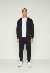Moleton preto com fecho éclair sobre uma t-shirt branca, combinado com calças pretas ajustadas e sapatilhas brancas. Design simples sem padrões notáveis.