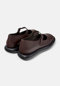 Zapatos de cuero marrón con punta redonda, diseño recortado y una suela acolchada negra. Presentan una correa decorativa con un detalle de lazo.