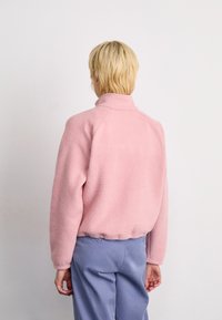Persona con capelli biondi corti che indossa una giacca in pile rosa chiaro e pantaloni blu, in piedi di fronte a una parete bianca uniforme.