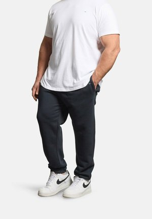 Mann trägt ein schlichtes weißes T-Shirt, schwarze Jogginghose und weiße Nike-Sneaker mit schwarzem Swoosh, steht mit einer Hand in der Tasche.