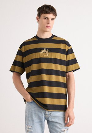 Striped t-shirt in yellow and black fabric, featuring a sun graphic on the chest. Short sleeves and relaxed fit. Worn with light blue distressed jeans.