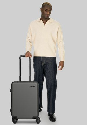 Joven con suéter crema y jeans oscuros sosteniendo una maleta gris con ruedas y mango extendido sobre un fondo gris liso.