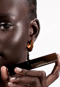 Corrector Luminous Lift 24HR de MAC Studio Radiance en un tubo oscuro. La mano presentada tiene una manicura beige claro, sosteniendo el tubo cerca de la cara de una modelo.
