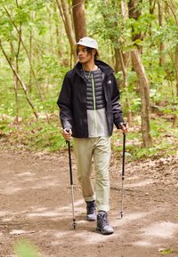 Nuori mies, jolla on hattu ja vaellusjalkineet, kävelee vaelluskeppien kanssa metsäreitillä, jota ympäröivät vihreät puut.