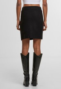 Černá pletená sukně s žebrovaným pasem, přiléhavým střihem a délky do středu stehna, v kombinaci s vysokými černými koženými botami s špičatým vrchem.