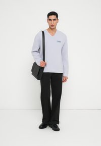 Violets krāsas adīta džemperis ar V-veida kaklu un logotipa uzšuvumu, kombinēts ar melnām biksēm un melnu pleca somu. Gluda tekstūra un brīva piegriezuma.