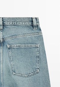 Lichtblauwe spijkerbroek die de achterzak, riemlussen en stikdetails toont op een effen witte achtergrond.