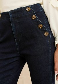 Temno modri jeans hlače z visokim pasom in edinstvenim stranskim gumbom, ki ima štiri okrogle, zlate gumbe. Gladka tekstura.
