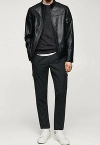 Giacca di pelle nera con zip frontale, abbinata a un maglione nero spesso, pantaloni scuri con tasche e sneakers bianche.