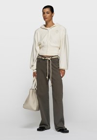 Frau trägt einen kurzen cremefarbenen Hoodie, locker sitzende graue Hose mit Kordelgürtel, hält eine strukturierte cremefarbene Handtasche und trägt schwarze Schuhe vor einem schlichten Hintergrund.