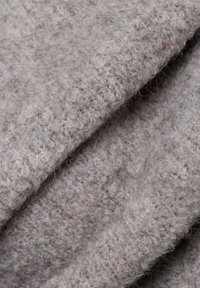 Strukturiertes Wollgewebe in hellem Beige, das ein geschlungenes Strickmuster und eine weiche, flauschige Oberfläche aufweist. Zeigt gefaltete Schichten mit leichtem Glanz.