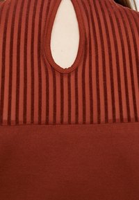 Gros plan sur un haut couleur rouille avec des rayures verticales transparentes et une petite ouverture en forme de goutte près du décolleté.