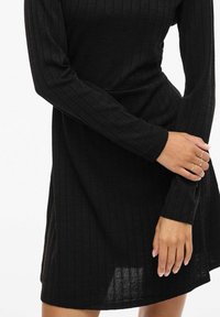 Robe noire à manches longues côtelée avec une coupe décontractée. Le tissu semble doux, présentant des rayures verticales et un léger éclat. Longueur mi-cuisse.