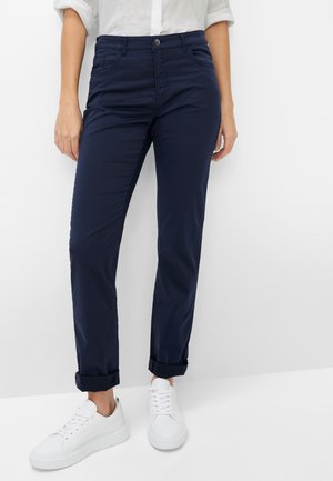 Femme portant un pantalon bleu marine retroussé, des baskets blanches et une chemise blanche à manches longues, debout devant un fond blanc uni.