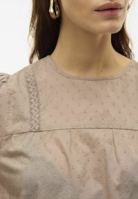 Blusa beige chiaro con pois testurizzati, caratterizzata da dettagli in pizzo lungo la cucitura della spalla e un collo rotondo. Tessuto morbido e liscio.