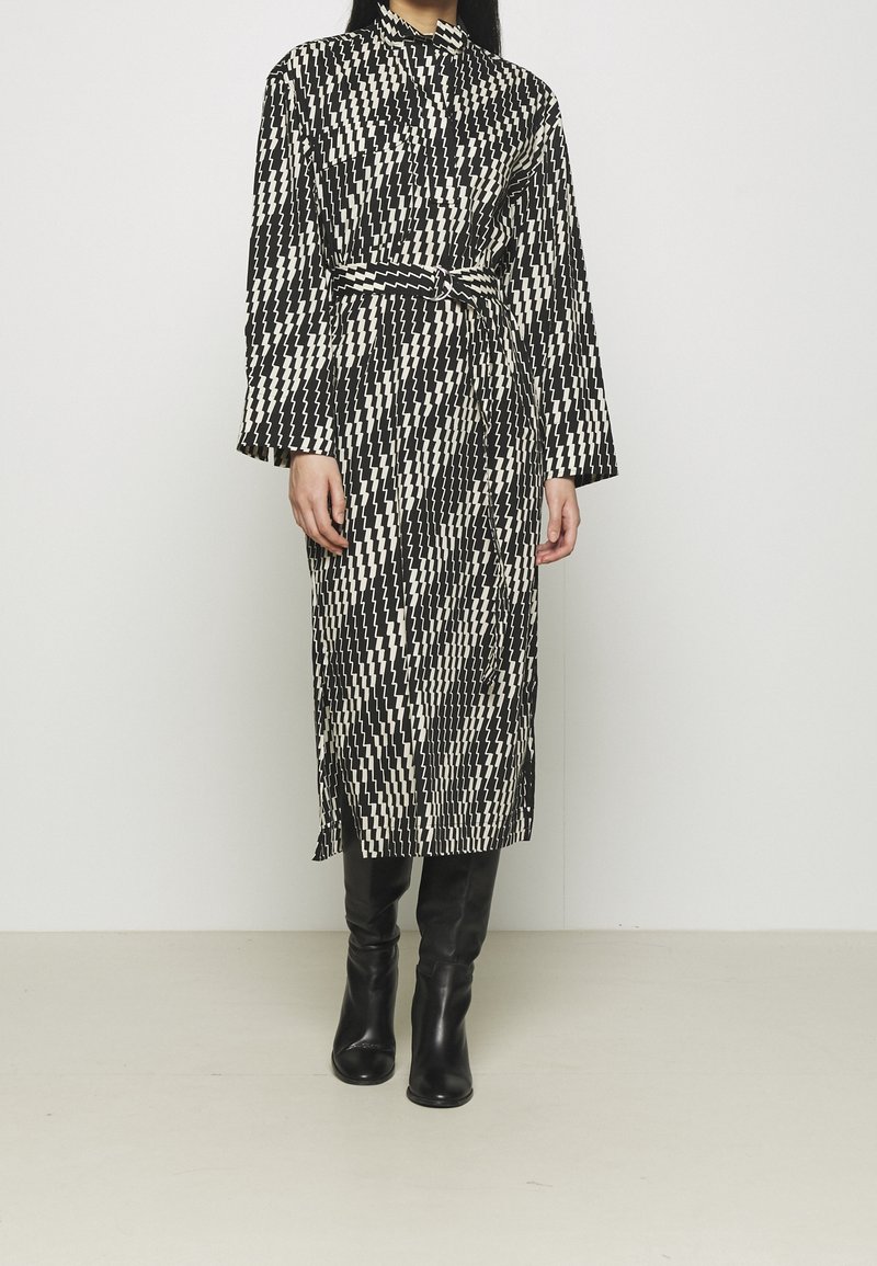 Personne portant une robe midi ceinturée à motif zigzag noir et blanc, manches longues et bottes noires montantes, debout sur un sol uni.