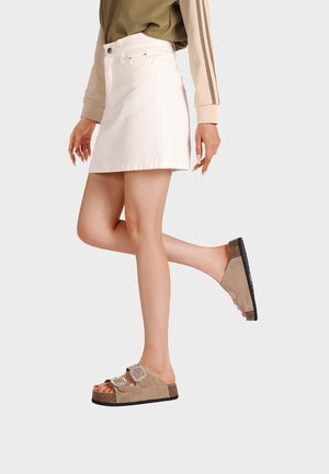 Donna che indossa un top beige a maniche lunghe e una minigonna bianca solleva una gamba, indossando sandali in pelle scamosciata beige con fibbie argento.