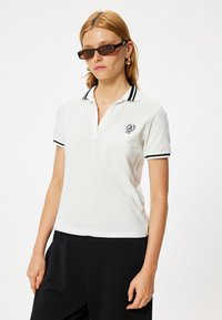Witte poloshirt met een zwart-wit gestreepte kraag, logo op de linkerborst en korte mouwen, gecombineerd met zwarte broeken.