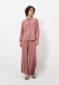 Lässiges, puderrosa Lounge-Set aus glattem Stoff; Hemd mit Knöpfen und Brusttasche, weite Hosen und Absatzsandalen.