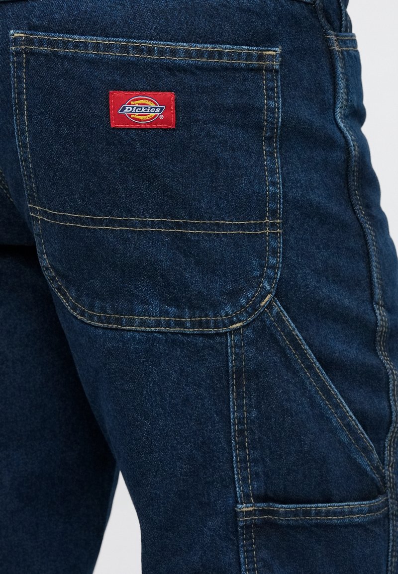 Dark blue denim pants featuring a back patch pocket with a red and yellow Dickies logo. Contrast stitching and durable material visible.