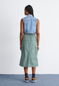 Vesta din denim, de culoare albastră deschisă, fără mâneci, asociată cu o fustă verde, până la genunchi, cu buzunare laterale. Modelul poartă șosete gri și adidași cu accente portocalii.