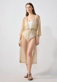 Áttetsző aranybársony kimonó stílusú felső, háromnegyedes ujjakkal és derékban megkötős. Könnyű, texturált anyagból készült, áramló kialakítással.