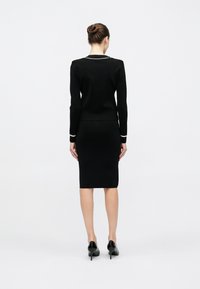 Μαύρο πλεκτό φόρεμα με εφαρμοστή σιλουέτα, με λευκή επένδυση στον γιακά και στις μανσέτες. Ομαλή υφή, μήκος μέχρι το γόνατο, σε συνδυασμό με μαύρες γόβες.