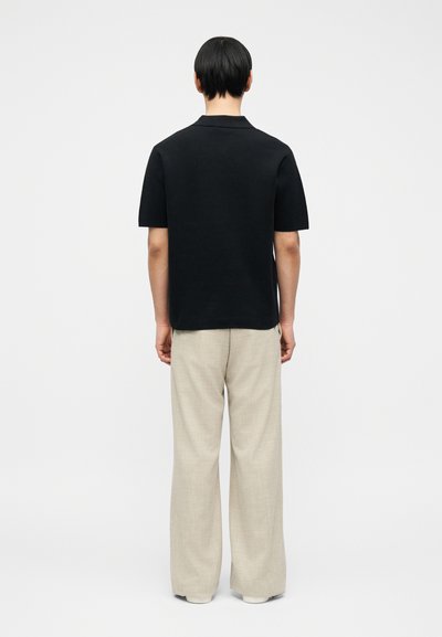 Άτομο με κοντά μαύρα μαλλιά που φοράει μαύρο κοντομάνικο πουκάμισο και άνετο μπεζ παντελόνι, στέκεται με το πρόσωπο στραμμένο προς ένα απλό λευκό φόντο.