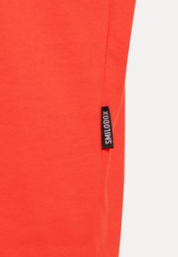 Tessuto arancione brillante con un piccolo etichetta nera con la scritta "SMILODOX" cucita in una cucitura sul lato destro.