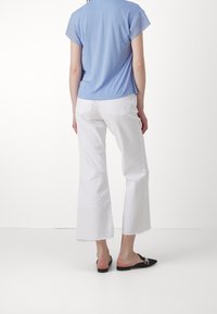 Personne portant un haut bleu clair à manches courtes, un pantalon blanc ample court et des chaussures noires sans talons avec un détail décoratif, debout sur fond blanc.