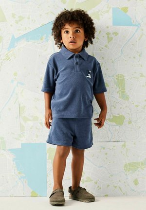 Jong kind met krullend haar, gekleed in een blauw poloshirt en bijpassende korte broek, staande tegen een muur bedekt met een stadskaart.