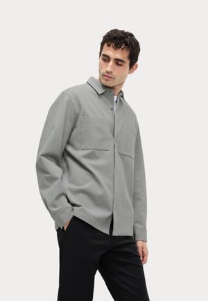 Camisa gris de manga larga con cuello, que presenta dos bolsillos delanteros y un corte holgado, hecha de tejido texturizado. Se lleva con pantalones negros.