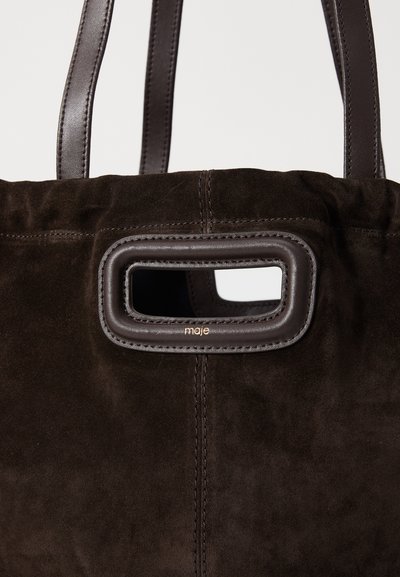 Brown suede tote bag with smooth leather accents, featuring a rectangular cutout handle and gold logo detail near the top.