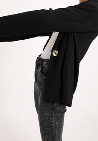 Person som bär en svart kavaj med en stor ljusbrun knapp över en vit skjorta och urtvättade svarta jeans, med armen sträckt rakt fram.