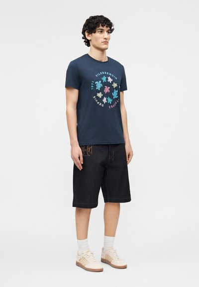 Νεαρός άνδρας που φοράει μπλε σκούρο t-shirt με σχέδιο χελώνας, μαύρο σορτς μέχρι το γόνατο με επιδιορθώσεις, λευκές κάλτσες και μπεζ αθλητικά παπούτσια.