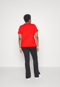 Person med kort, krøllet hår iført en rød t-shirt, sorte bukser og hvide sneakers, stående foran en ensfarvet lys grå baggrund.