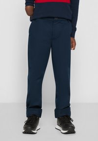 Marineblå bukser med lige snit, prydet med knaplukning og opfoldede bukseben, parret med sorte sneakers. Glat stofstruktur.