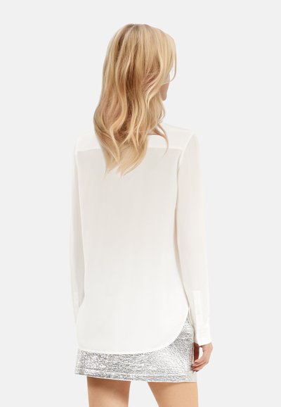 Weiße Bluse mit langen Ärmeln aus weichem Stoff, geschwungenem Saum und kontrastierendem, durchsichtigem Schulterbereich, kombiniert mit einem silbernen, strukturierten Rock.