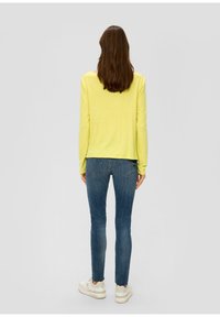 Geel languit shirt met een relaxte fit, gecombineerd met blauwe slimfit jeans en witte sneakers. De textuur lijkt zacht; het ontwerp is eenvoudig.