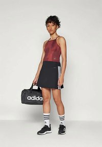 Femme en débardeur bordeaux et jupe noire avec des rayures blanches, tenant un sac de sport noir Adidas, portant des chaussettes blanches Adidas et des baskets noires.