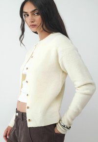Cremfarbener Strickcardigan mit fünf Holzknöpfen, rundem Ausschnitt und gerippten Bündchen. Trägt man über einem weißen Top und kombiniert mit braunen Hosen.
