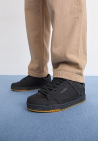 Personne portant un pantalon beige et des baskets Globe noires à semelles gomme, debout sur un sol recouvert d'un tapis bleu.