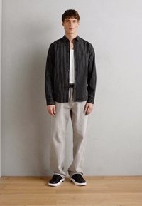Camisa de botão às riscas preto e branco, top branco, calças cinza claro e ténis pretos com solas brancas. De pé contra um fundo simples.