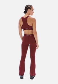 Bordó aktív szett, amely tartalmaz egy pántos sportmelltartót és bővülő szárú leggingset, rugalmas anyagból készült, a derékon logós szalaggal.