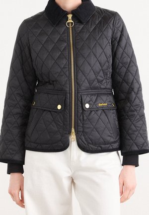 Schwarze gesteppte Jacke mit goldfarbenem Reißverschluss und Knöpfen, zwei vordere Klappentaschen, getragen über weißen Hosen, Markenname „Barbour“ sichtbar auf der Tasche.