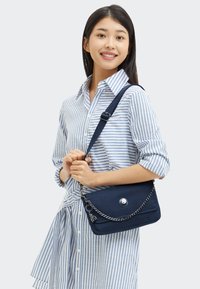 Giovane donna sorridente, che indossa un vestito a righe blu e bianche, mentre tiene una borsa a tracolla blu navy con dettagli in catena.