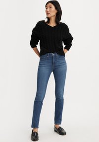 Maglione nero a treccia con scollatura a V, abbinato a jeans skinny blu a vita media e mocassini neri. Design semplice e strutturato, vestibilità elegante.