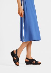 Sandali con zeppa neri in pelle con cinturini incrociati e fibbia alla caviglia, abbinati a un vestito blu con strisce bianche laterali.